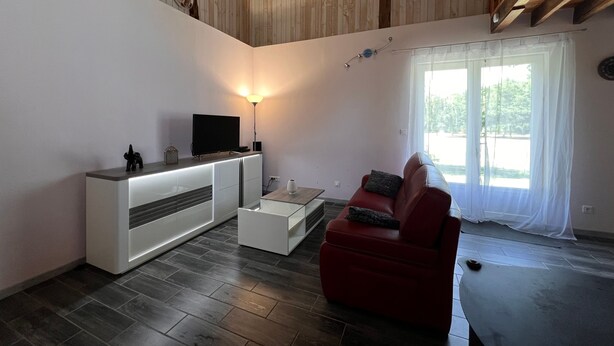 A modern living room with a flat-screen TV, a white entertainment unit, a red leather sofa, a glass coffee table, and a large window with sheer curtains.