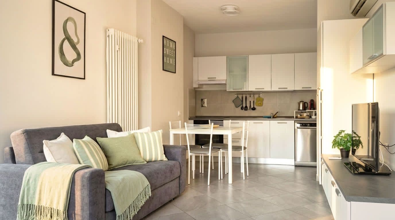 A modern living room with a grey sofa, a dining table with chairs, and a kitchen area with white cabinets and appliances.