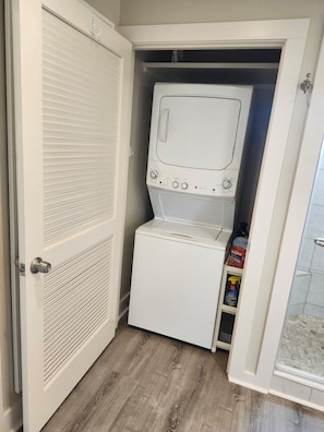 Un lavadero con lavadora y secadora de carga frontal, una puerta con diseño persiana y un estante con productos de limpieza.