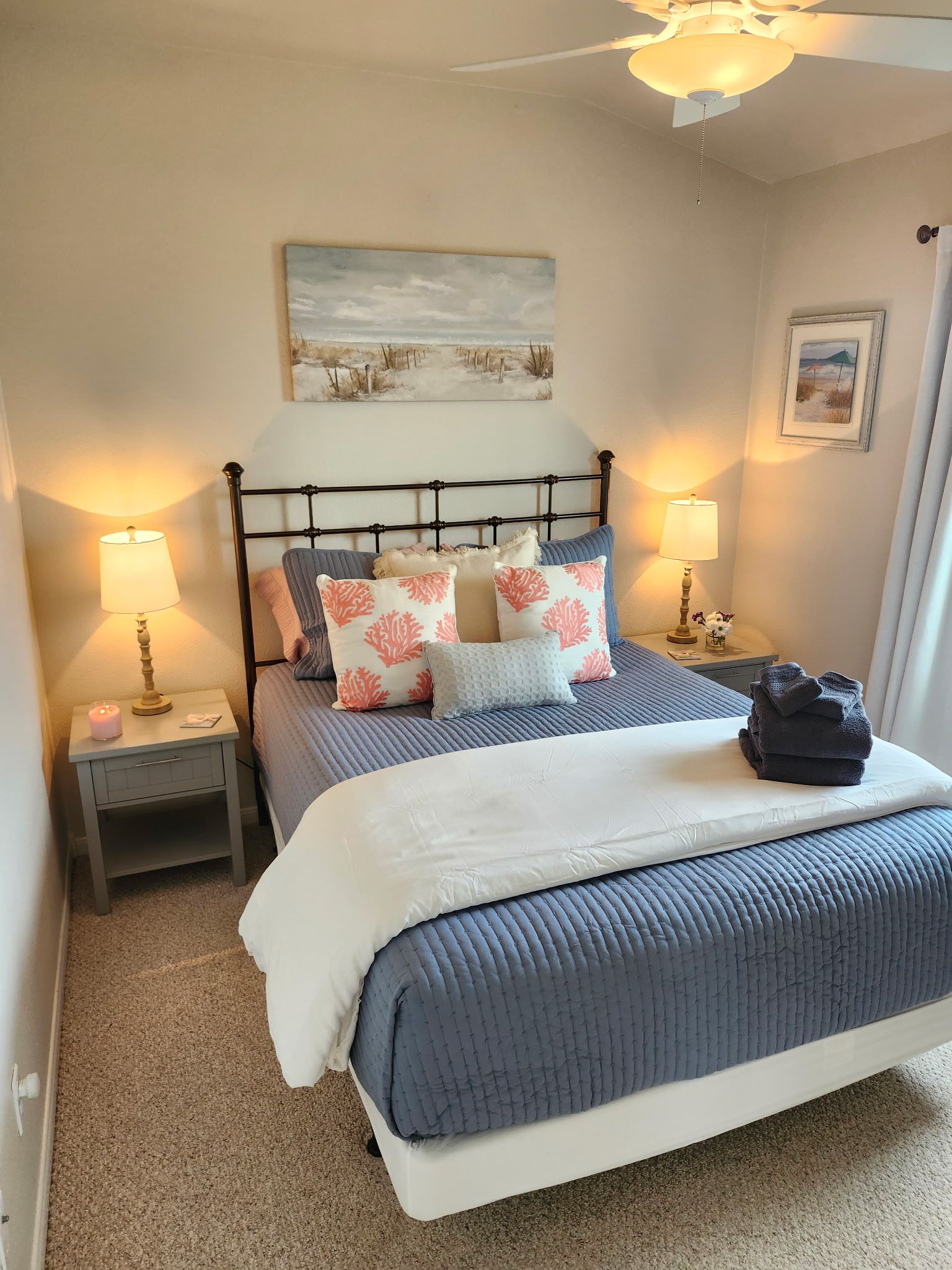 A neatly made bed with a blue quilt, a white pillow, and several decorative pillows with coral patterns. There are two bedside tables with lamps, a painting above the bed, and a framed picture on the wall.