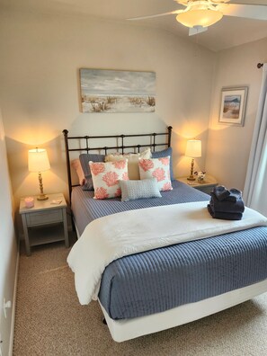 Een net opgemaakt bed met een blauwe quilt, een wit kussen en meerdere decoratieve kussens met koraalmotieven. Er zijn twee nachtkastjes met lampen, een schilderij boven het bed en een ingelijste foto aan de muur.