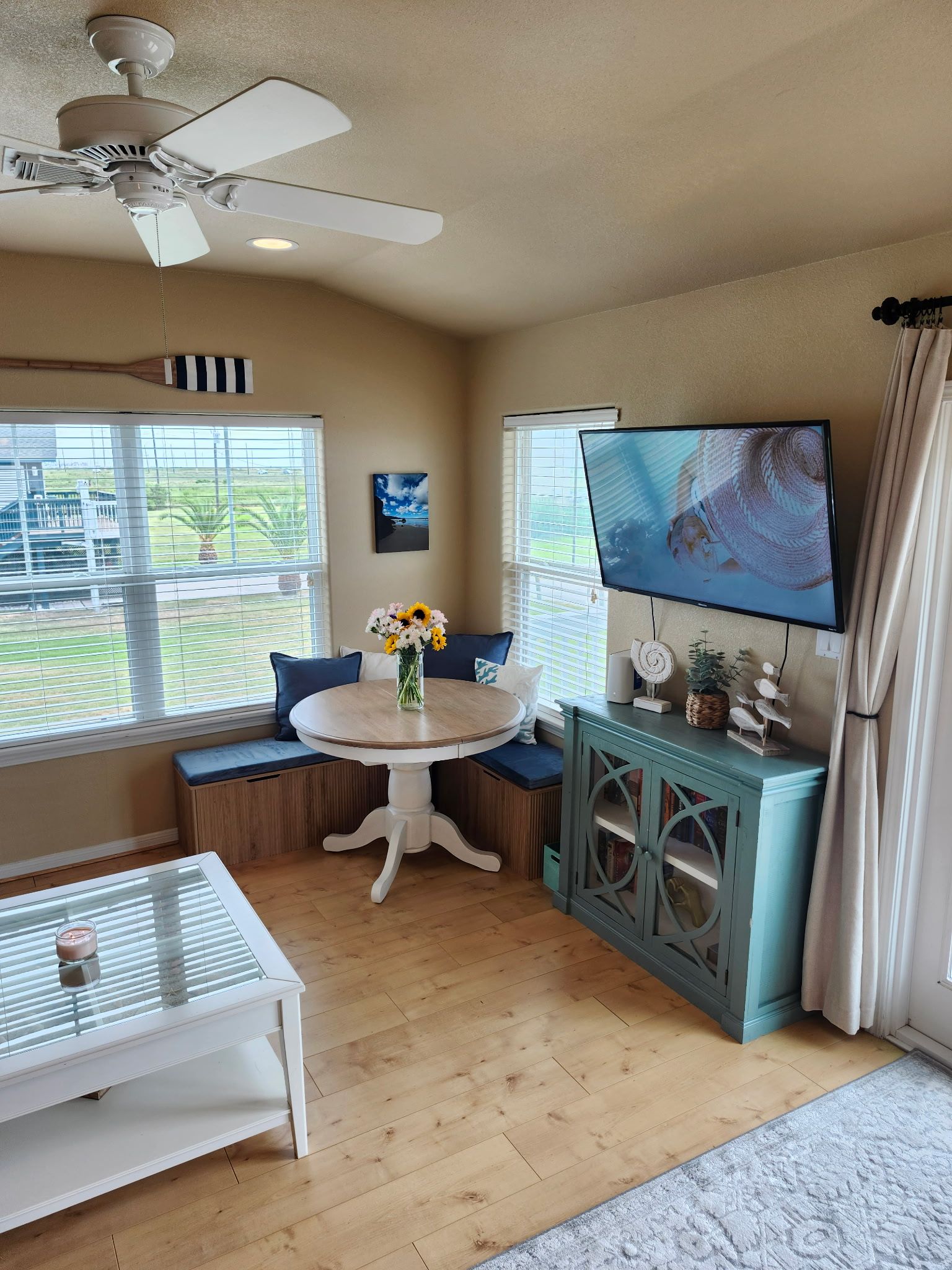 Una habitación con ventilador de techo, una mesa redonda con un florero, un televisor en la pared y una ventana con persianas.