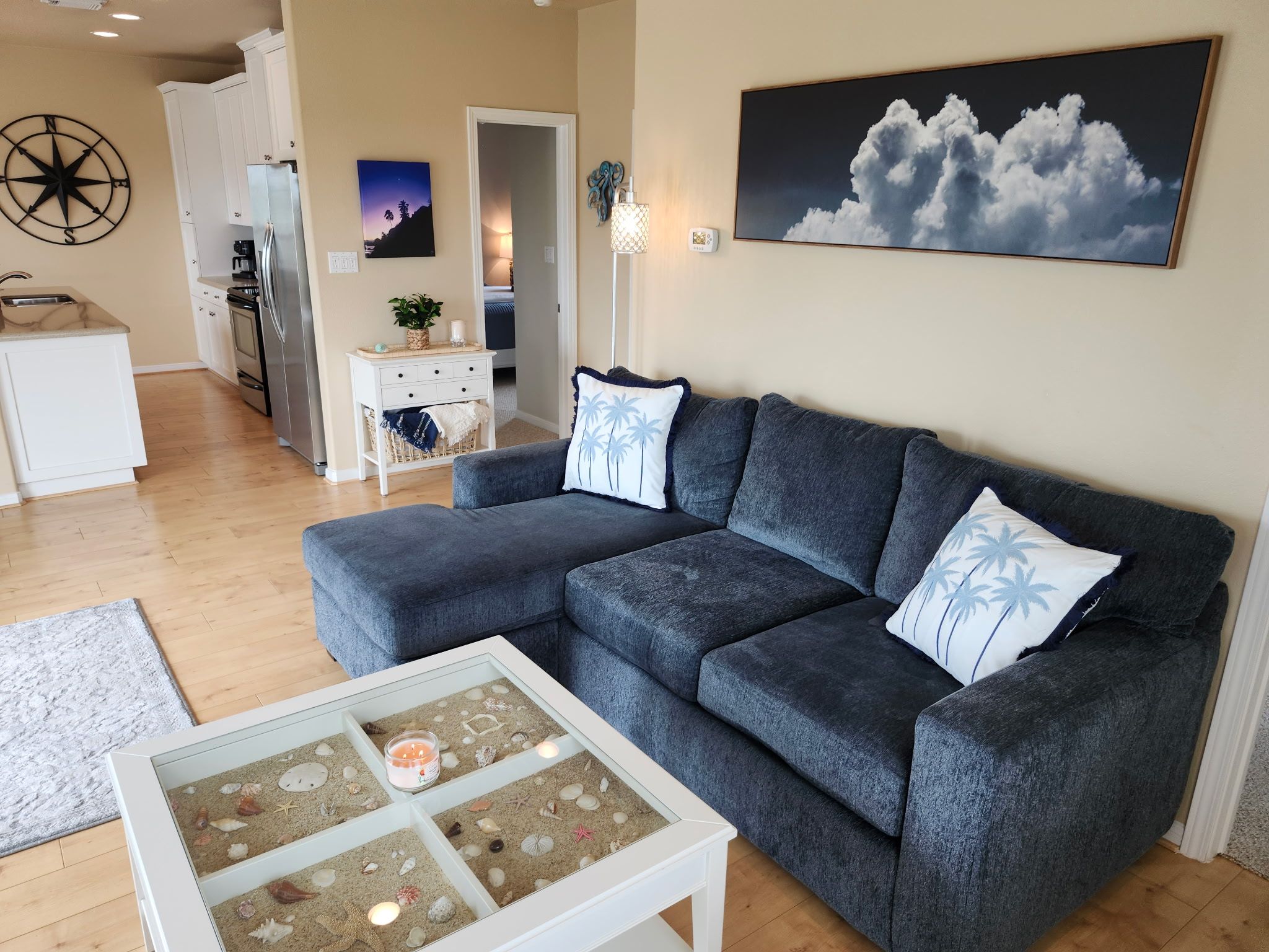 En stue med en grå sofa, et sofabord med et strandtema og et maleri af skyer på væggen.