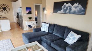 Uma sala de estar com um sofá cinza, uma mesa de centro com tema praiano e uma pintura de nuvens na parede.