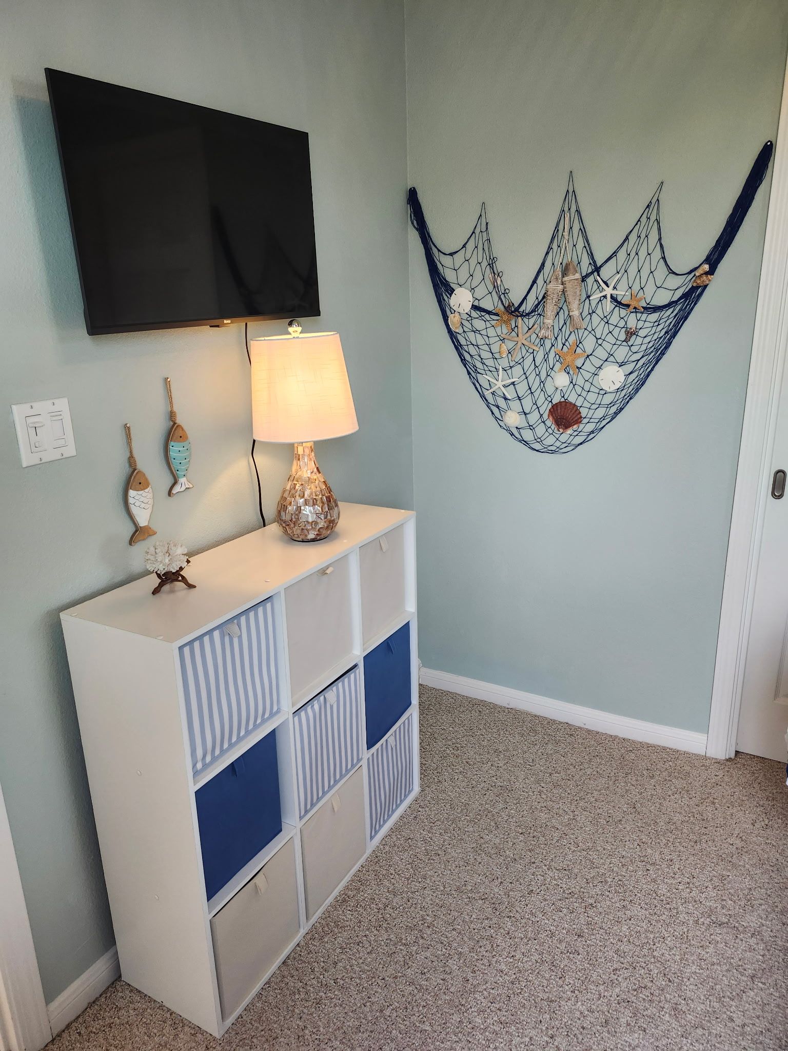 A room with a flat-screen TV mounted on the wall, a white console table with a lamp, and a decorative wall hanging.