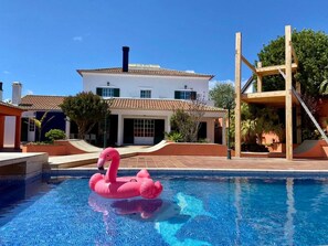 Una piscina con un flotador de flamenco rosa, un edificio blanco con techo de tejas y una estructura de madera a la derecha.