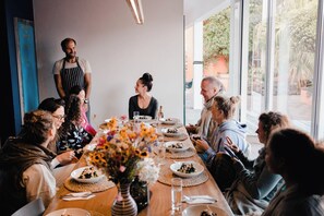 Un grupo de personas sentadas alrededor de una larga mesa de madera con platos de comida, en una habitación con ventanas amplias y una puerta azul.
