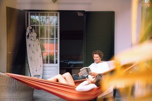 Una persona recostada en una hamaca leyendo un libro, con una tabla de surf apoyada contra una puerta.