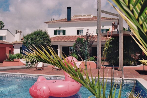 Una piscina con un flotador inflable rosa, una terraza de madera y un edificio con techo de tejas.