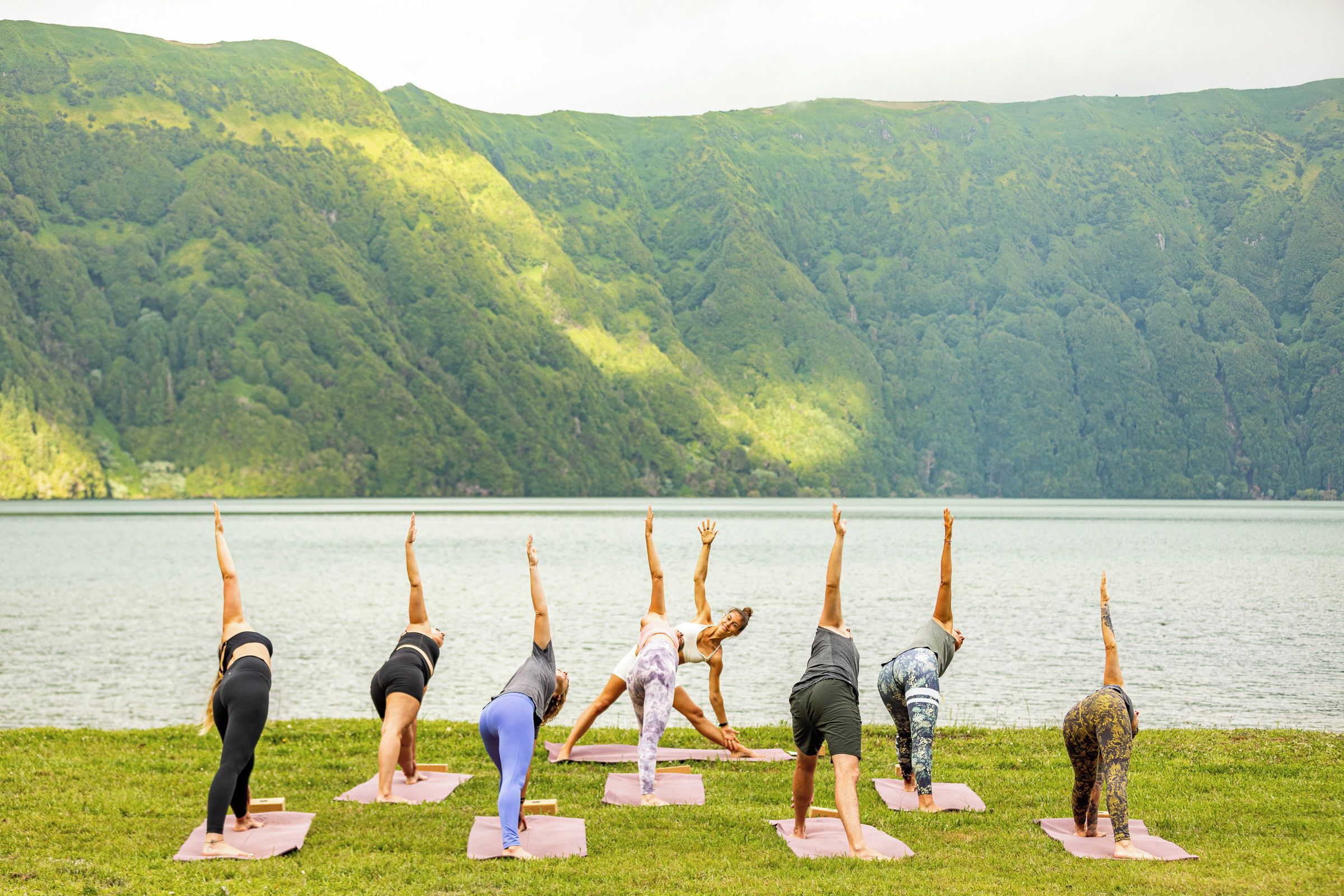 A group of people performing yoga on mats by a lake with a forested hill in the background.