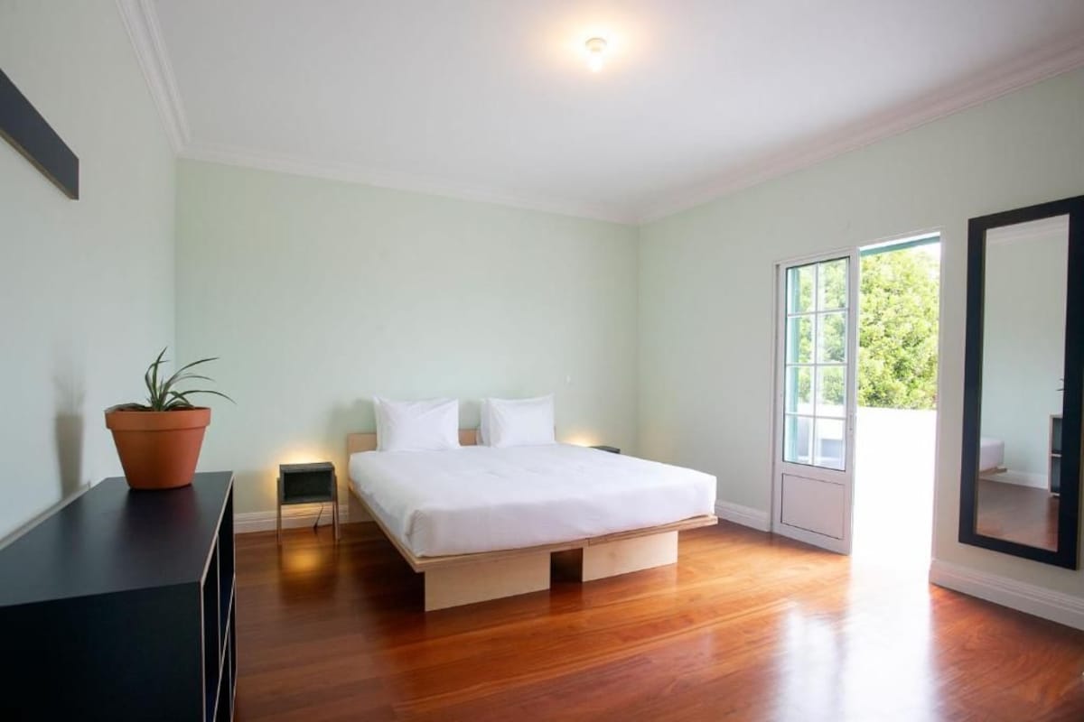 A bedroom with a bed, a potted plant, a small bedside table, and a large window.