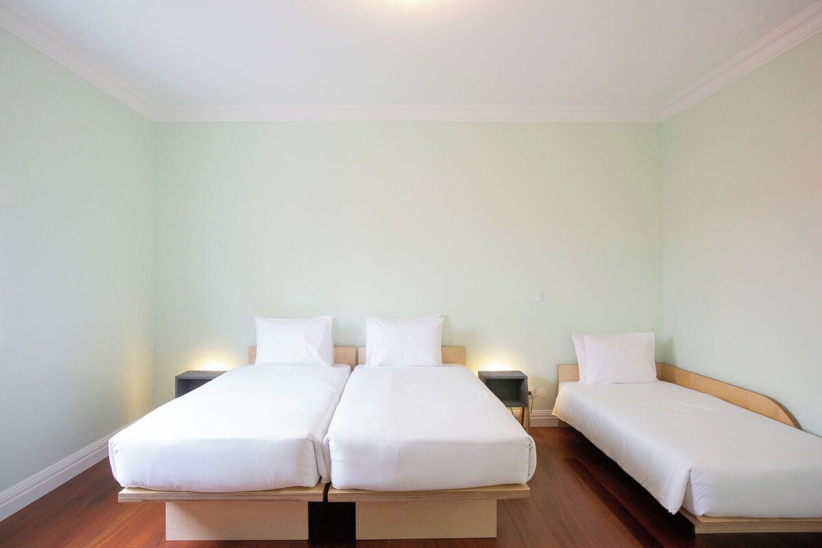 A hotel room with three single beds, each with a wooden headboard and white bedding. There are bedside tables with lamps.