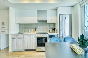 Eine moderne Küche mit weißen Schränken, einem Edelstahlbackofen, einer Mikrowelle und einem Kühlschrank. Ein Esstisch mit Stühlen und ein Topf mit einer Pflanze stehen am Fensterbrett.