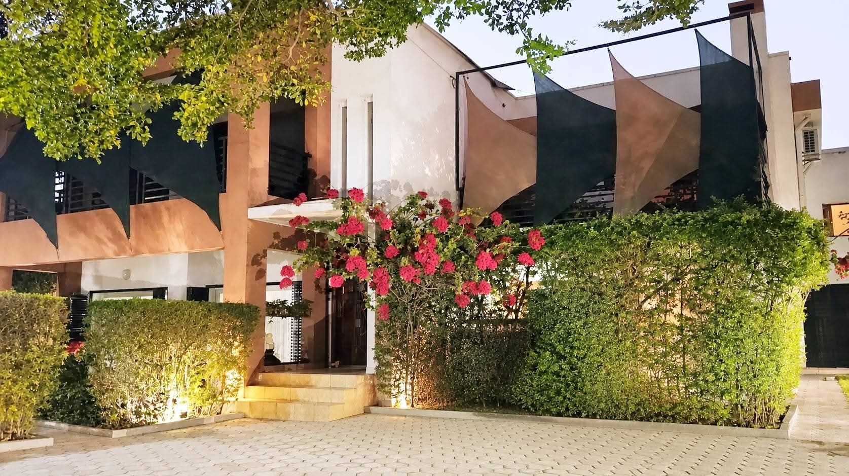 Un bâtiment doté d’une architecture moderne, avec un escalier menant à l’entrée, le tout entouré de haies bien entretenues et de plantes en fleurs.