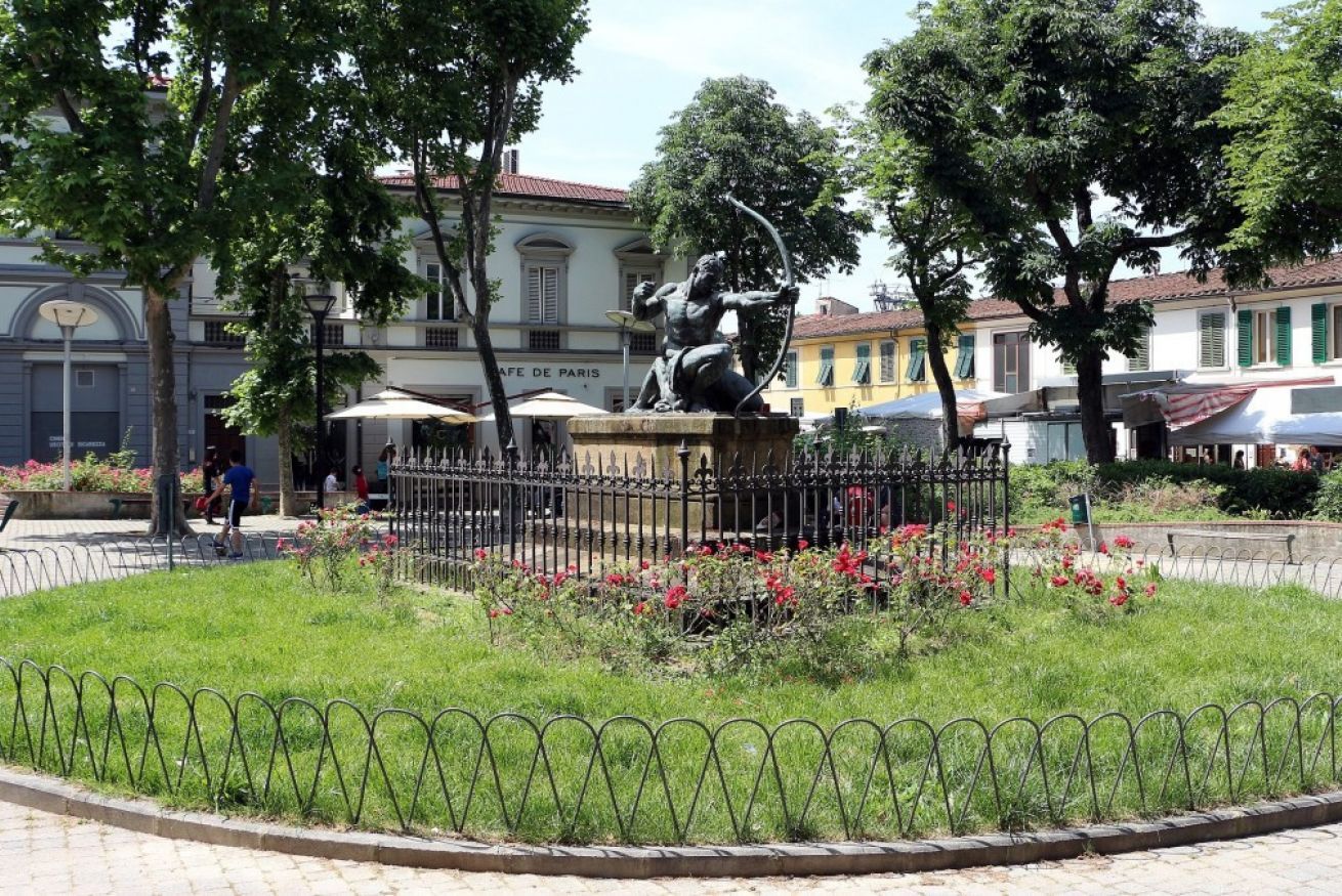 Une statue représentant une figure tenant un arc, placée sur une place publique, avec une clôture, des arbres et des bâtiments en arrière-plan.