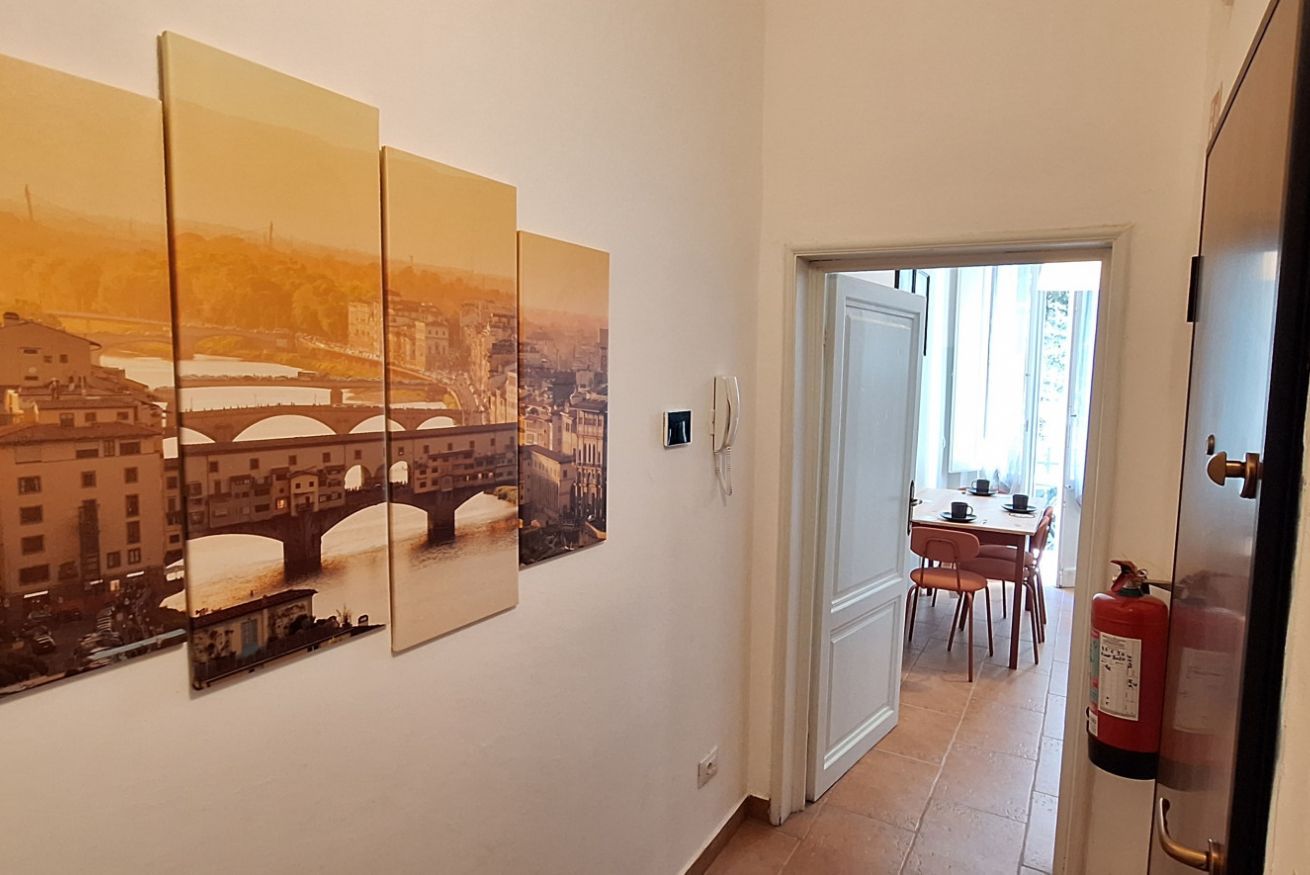 Un couloir orné d’un tableau représentant un paysage urbain, un coin repas avec des chaises et une table, et un extincteur fixé au mur.