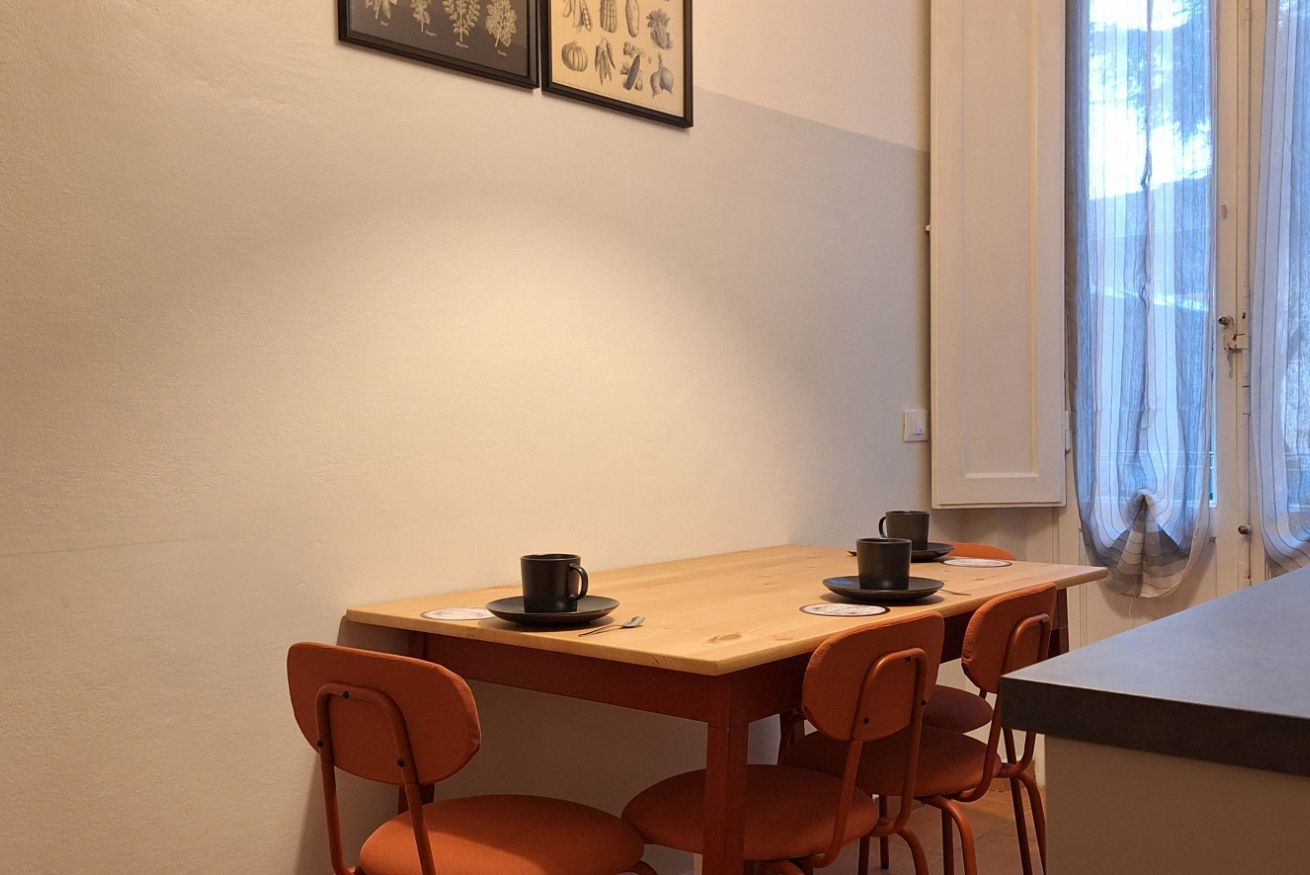 Un coin repas avec une table en bois et des chaises orange, une fenêtre avec des rideaux et, au mur, des reproductions encadrées de motifs botaniques.