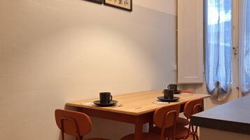Un coin repas avec une table en bois et des chaises orange, une fenêtre avec des rideaux et, au mur, des reproductions encadrées de motifs botaniques.