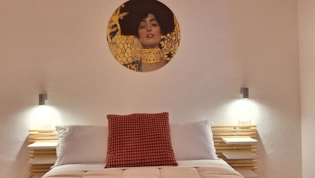Quarto com cama, duas mesinhas de cabeceira, luminária na parede e uma obra de arte circular sobre a cama.