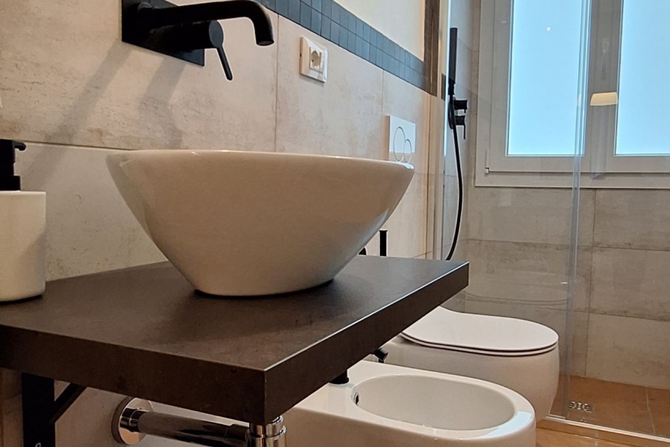 A bathroom with a large white bowl sink on a dark countertop, a white toilet, and a window with a view.