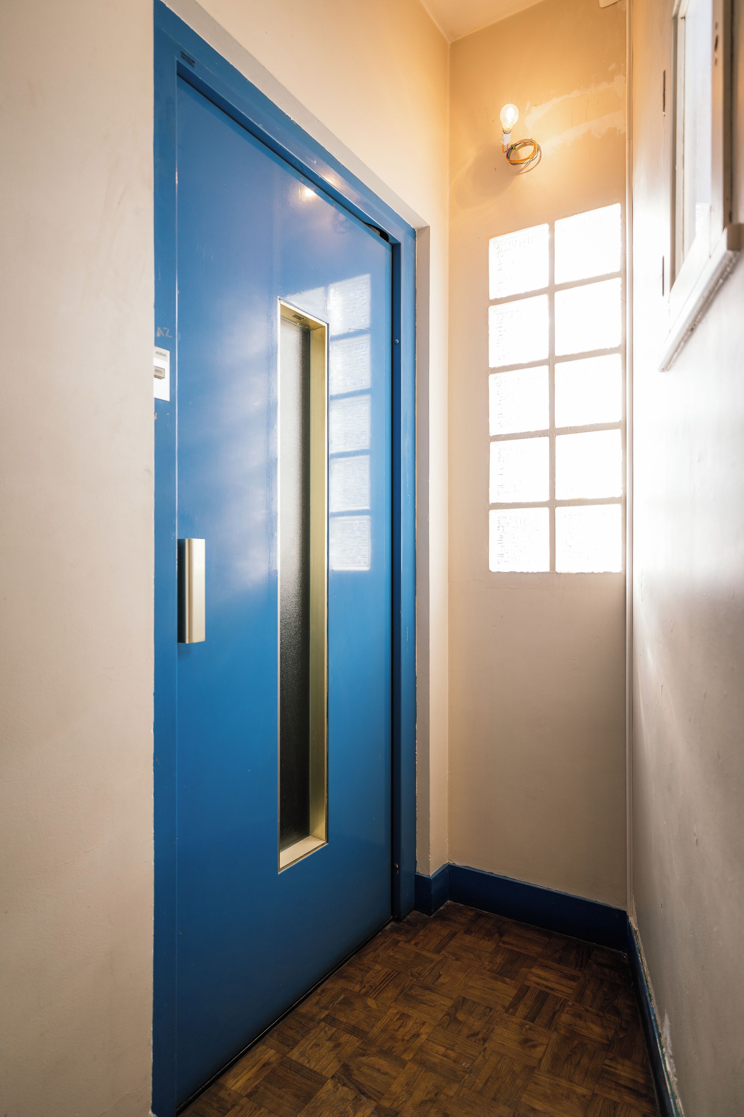 Een hal met een blauwe deur, een raam met zonwering en een houten vloer.
