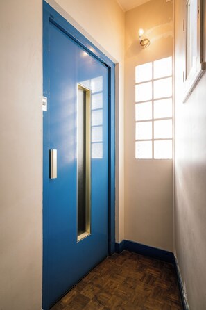 Een hal met een blauwe deur, een raam met zonwering en een houten vloer.