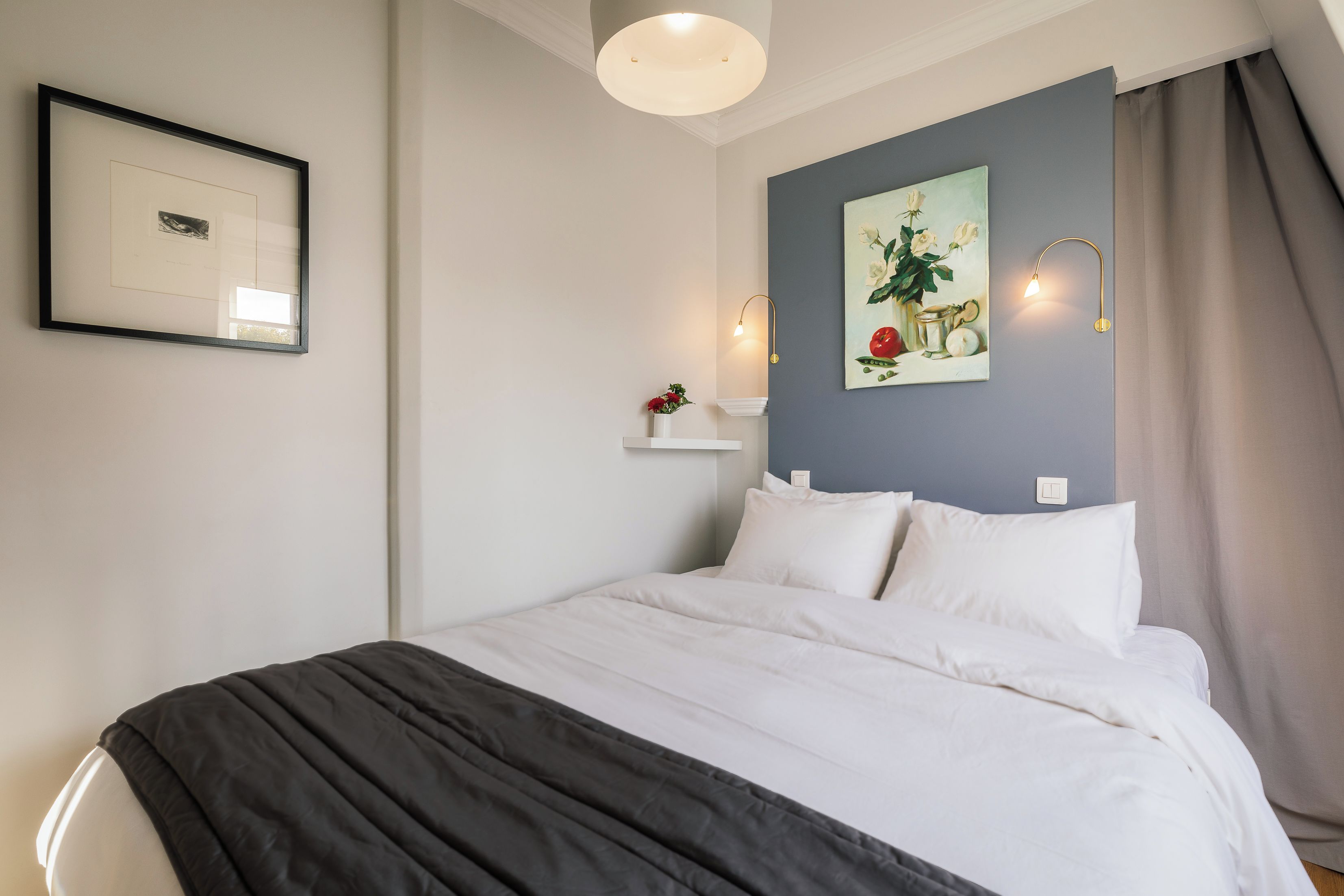 Un lit bien fait, avec du linge de lit blanc, un plaid gris foncé et un luminaire fixé au mur.