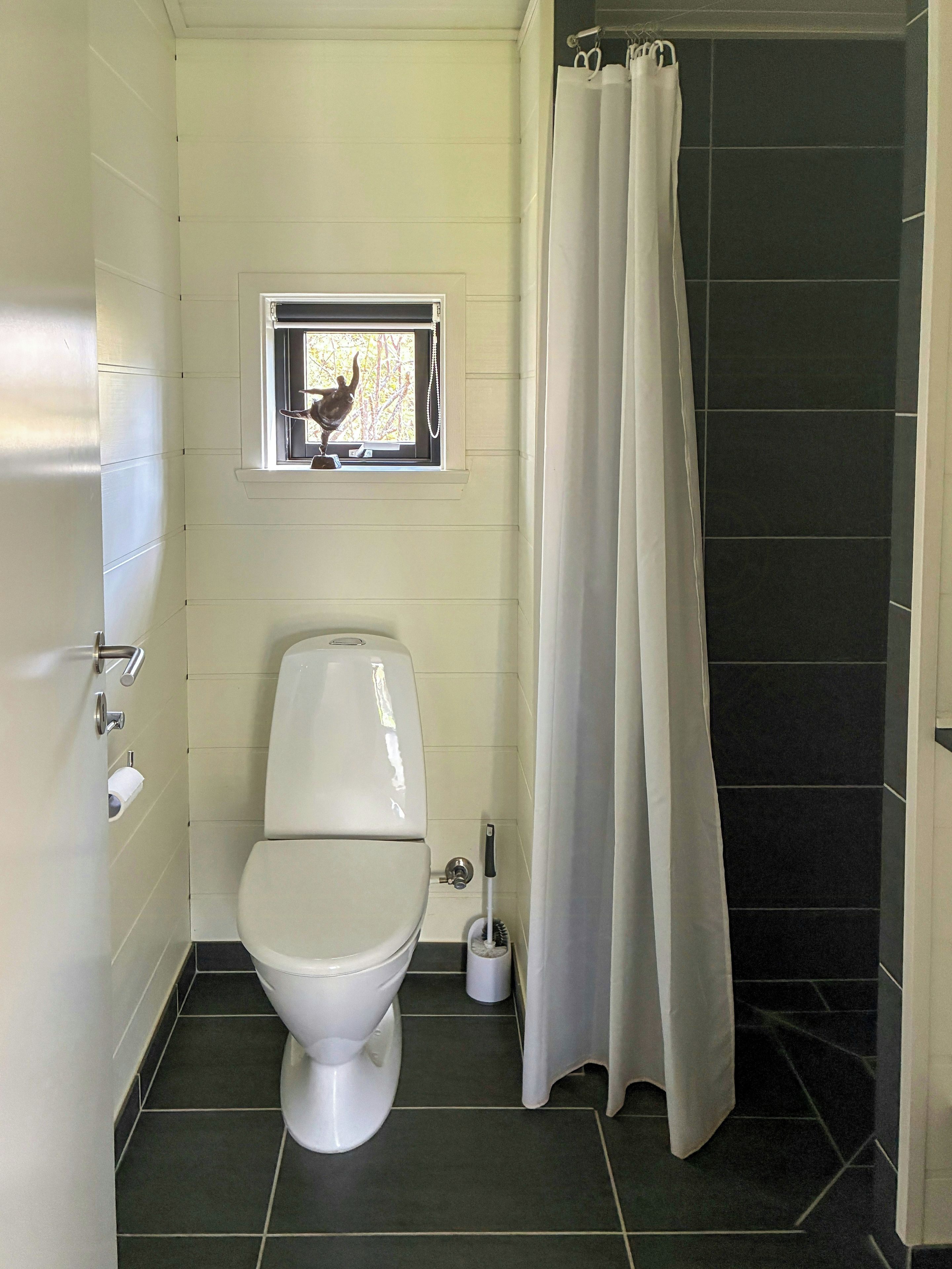 A bathroom with a toilet, a window, a shower curtain, and a toilet brush.