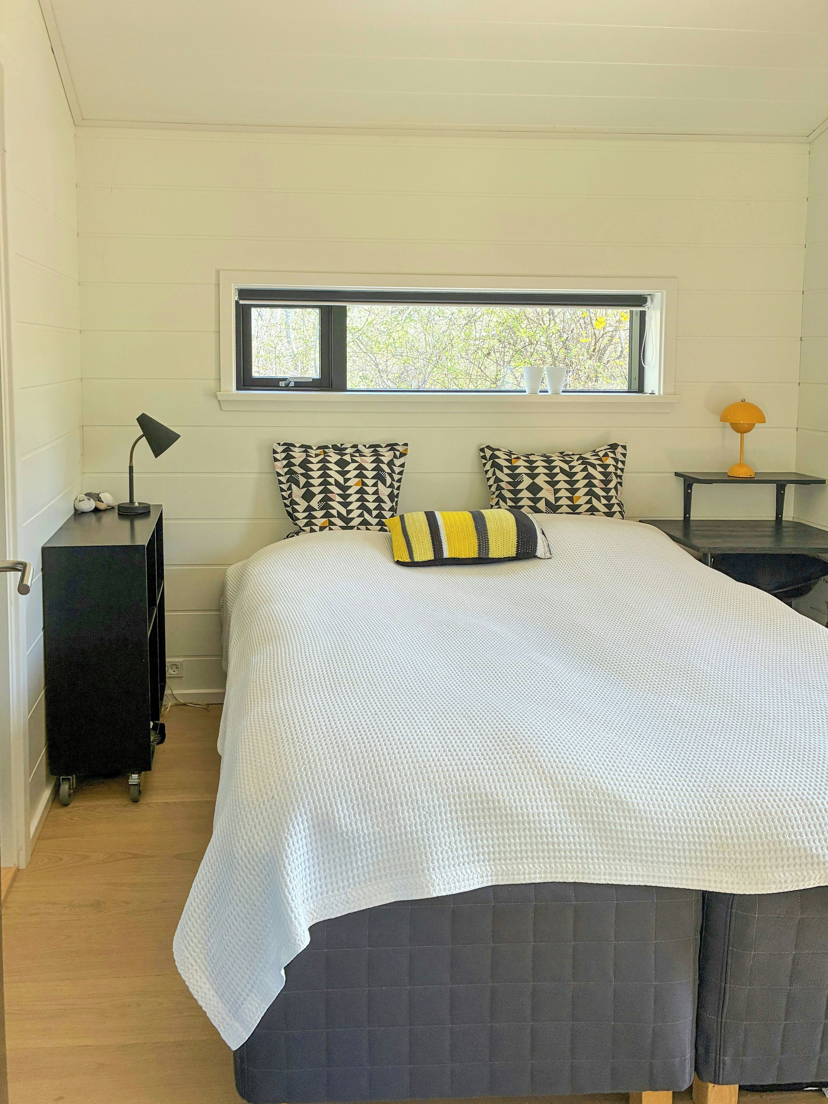A bedroom with a bed, pillows, a nightstand, a lamp, a window, and a view of trees.