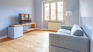 A modern living room with a grey sofa, a TV on a wooden stand, a radiator, and a window with blinds. - Luciano Boutique Apartment (Roma)