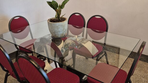 Una mesa con cubierta de vidrio, seis sillas rojas, una planta en maceta y dos cuadros en la pared.