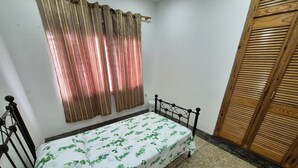 Una cama individual con estructura metálica, una colcha blanca con motivos de hojas verdes, una puerta de madera y una ventana con cortinas marrones.