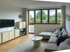 A living room with a flat-screen TV, a sofa, a coffee table, and a large window with a view of greenery. - Spacious flat by Amager Fælled and the harbour (København)