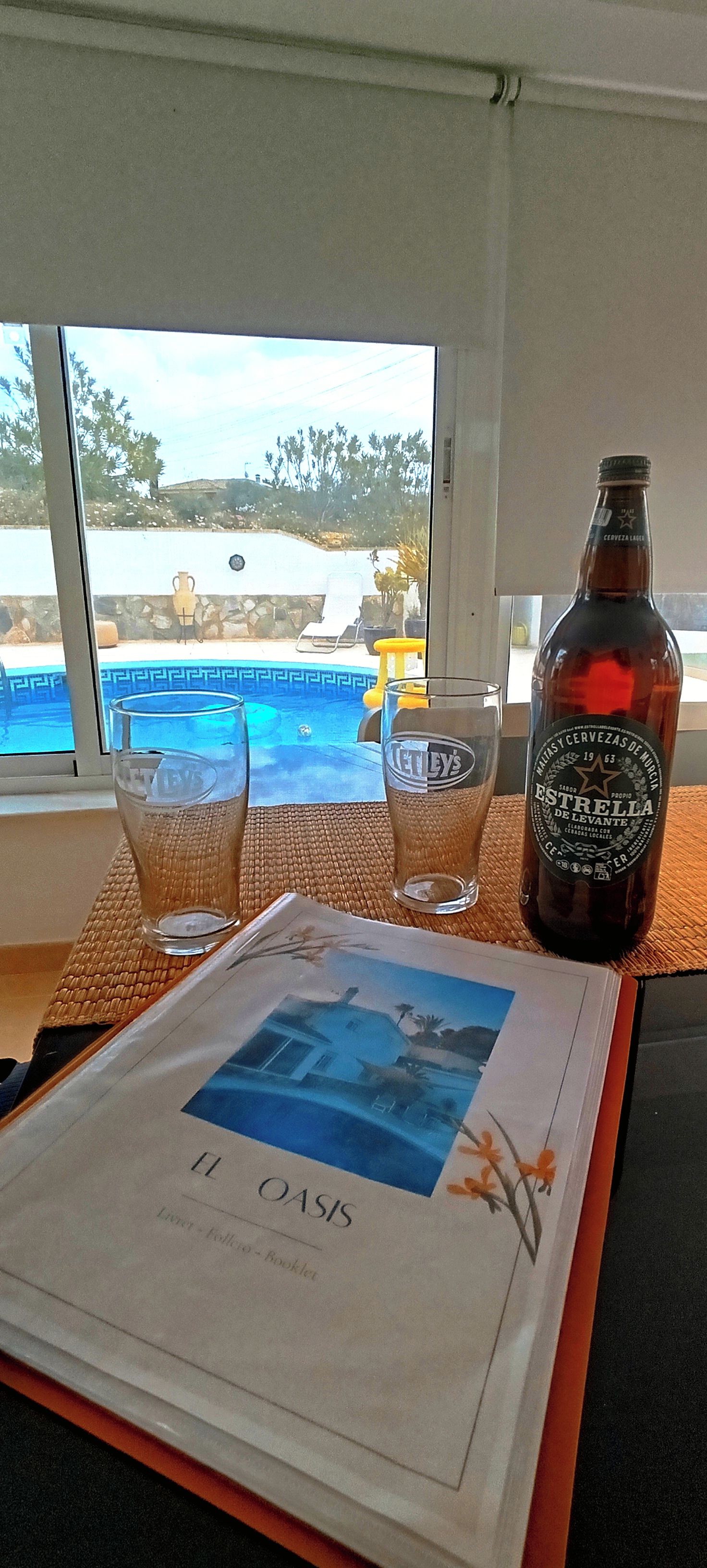 Een fles Estrella bier, twee glazen en een boekje op een tafel met uitzicht op een zwembad.