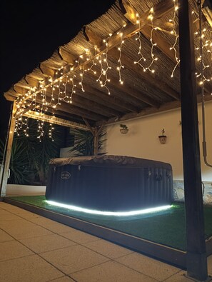 A hot tub under a wooden pergola with string lights, surrounded by a tiled patio and a small garden. - El Oasis 🌴 Private Home | Winter Spa & Summer Pool | Near Leana Thermal Baths (MC)