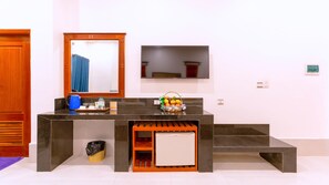 Habitación de hotel con puerta de madera, una encimera negra con hervidor, frutero, refrigerador pequeño y un cesto de basura.