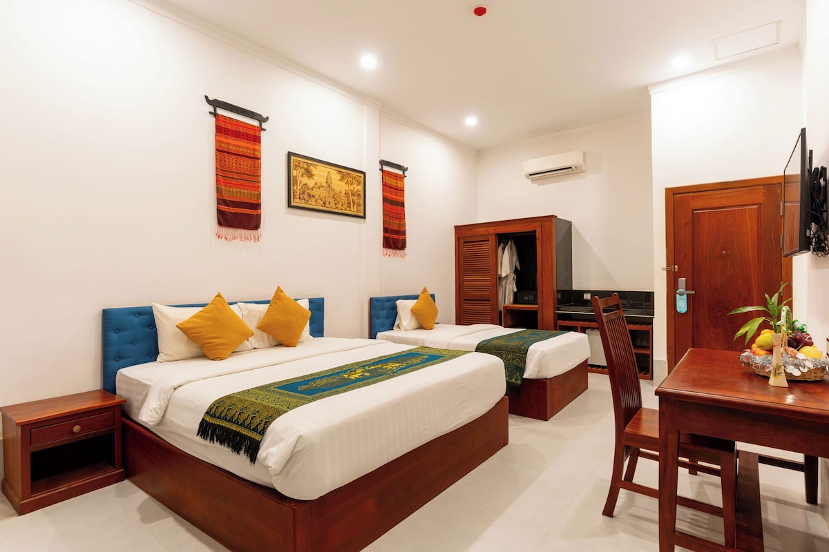 A hotel room with two beds, a wooden wardrobe, a desk with a chair, and a television.