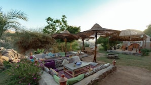 An outdoor seating area with thatched umbrellas, cushioned seating, and a stone wall. - Krouma Camp & Ecolodge (Aswan)