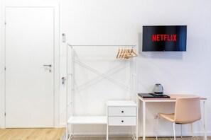 Una habitación con un escritorio blanco, una silla, un armario blanco, una percha para ropa y un televisor que muestra el logotipo de Netflix.