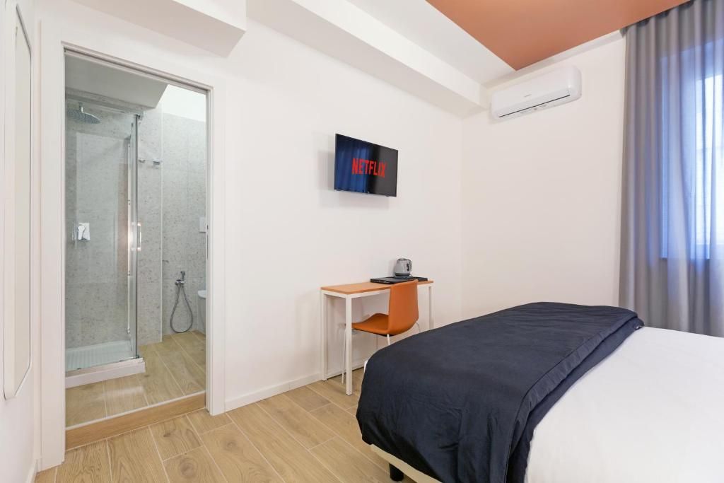 Quarto de hotel com cama, escrivaninha, cadeira, televisão e chuveiro.