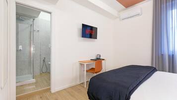 Quarto de hotel com cama, escrivaninha, cadeira, televisão e chuveiro.