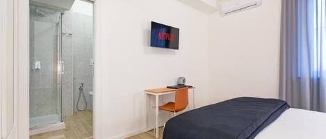 A hotel room with a bed, a desk, a chair, a TV, and a shower area.