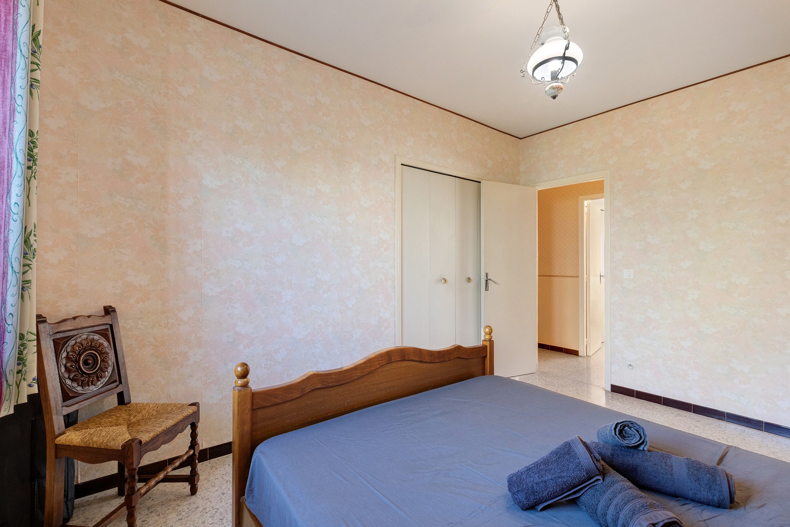 A bedroom with a bed, a chair, a wardrobe, and a door leading to another room.