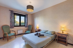 A bedroom with a bed, two chairs, a window with curtains, a bedside table with a lamp, and a wall clock.