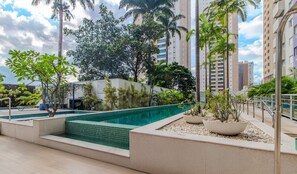 Una piscina con bordes de azulejos, rodeada de macetas con pequeños árboles y arbustos, y edificios altos al fondo.