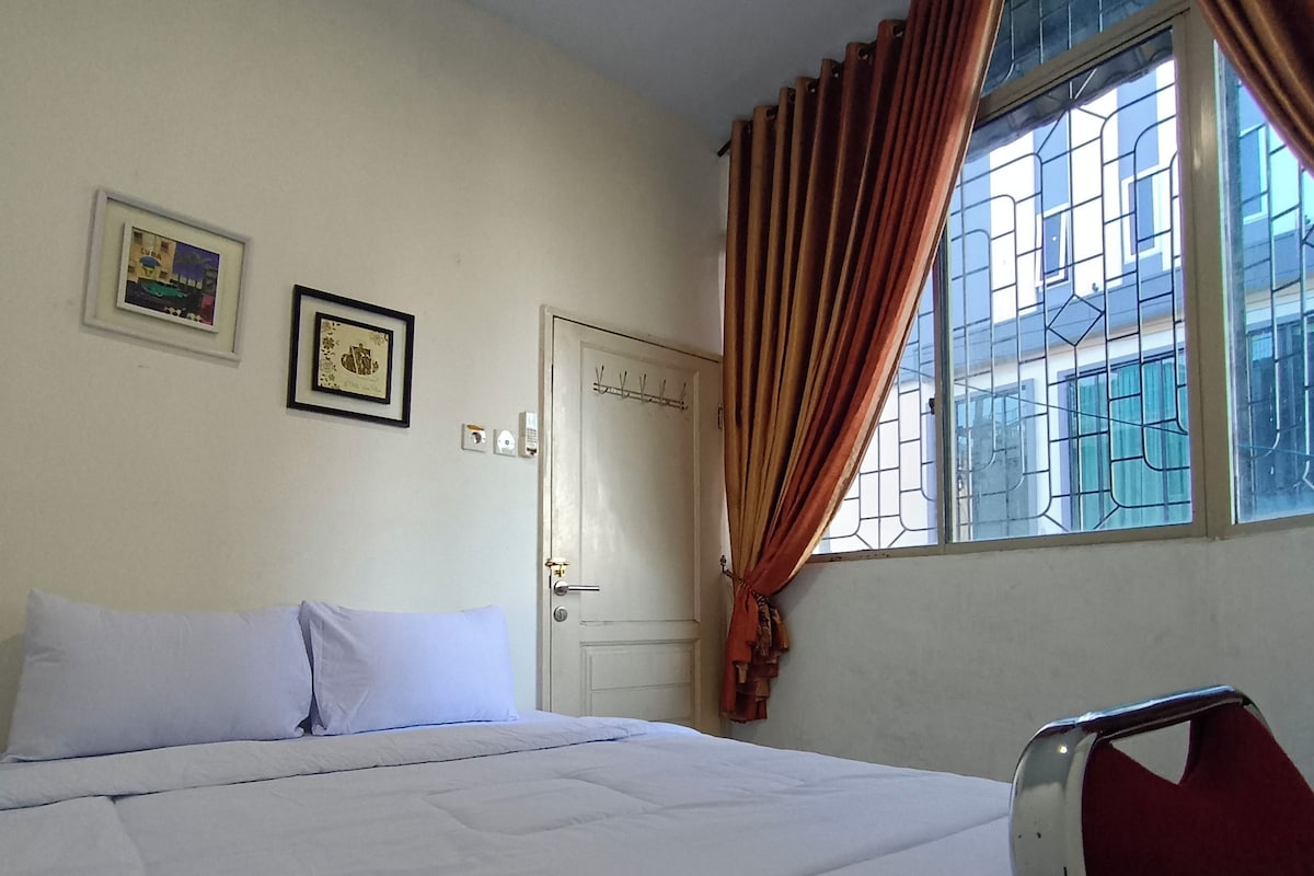 A room with a bed, a window with curtains, a door, and two framed pictures on the wall.