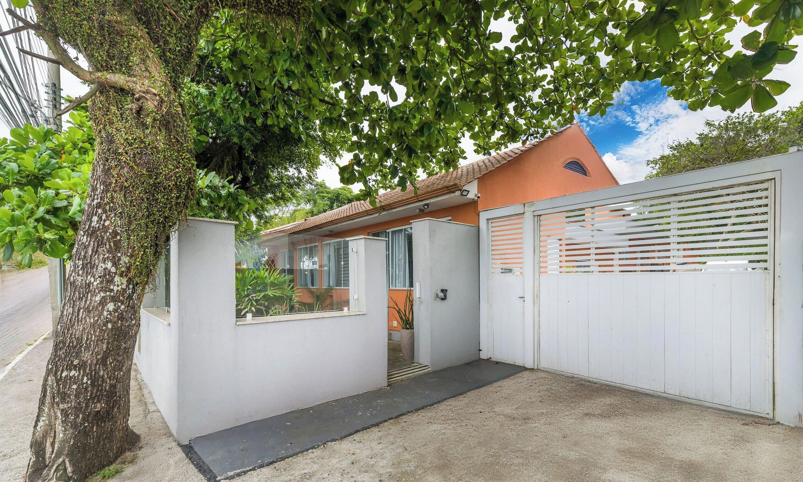 A gated residential property with a concrete driveway, a white wall with a window, and a tree with green leaves.