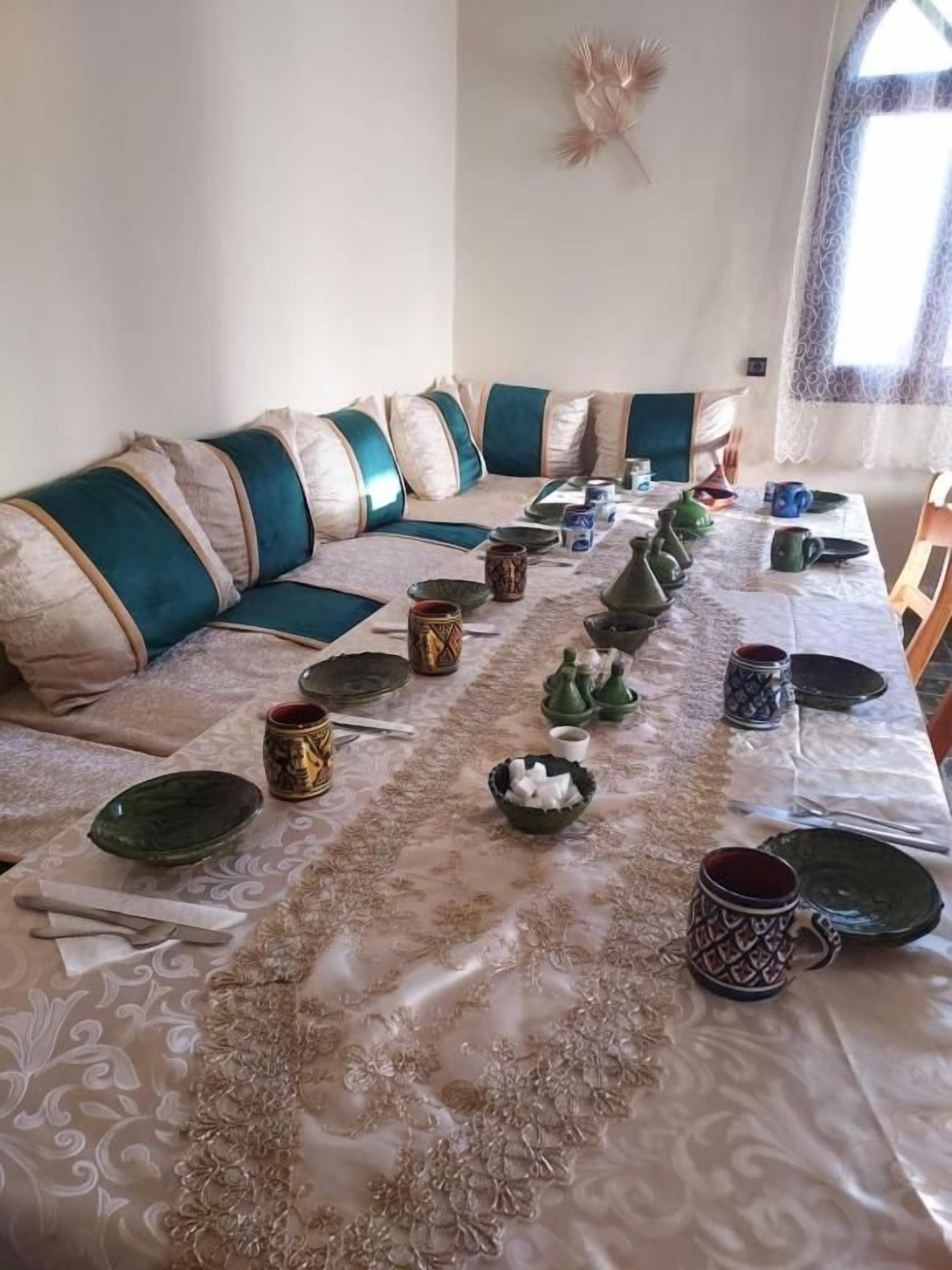 Un comedor con una mesa larga dispuesta para una comida, con mantel estampado, cojines verdes y beige, y diversos platos y cubiertos.