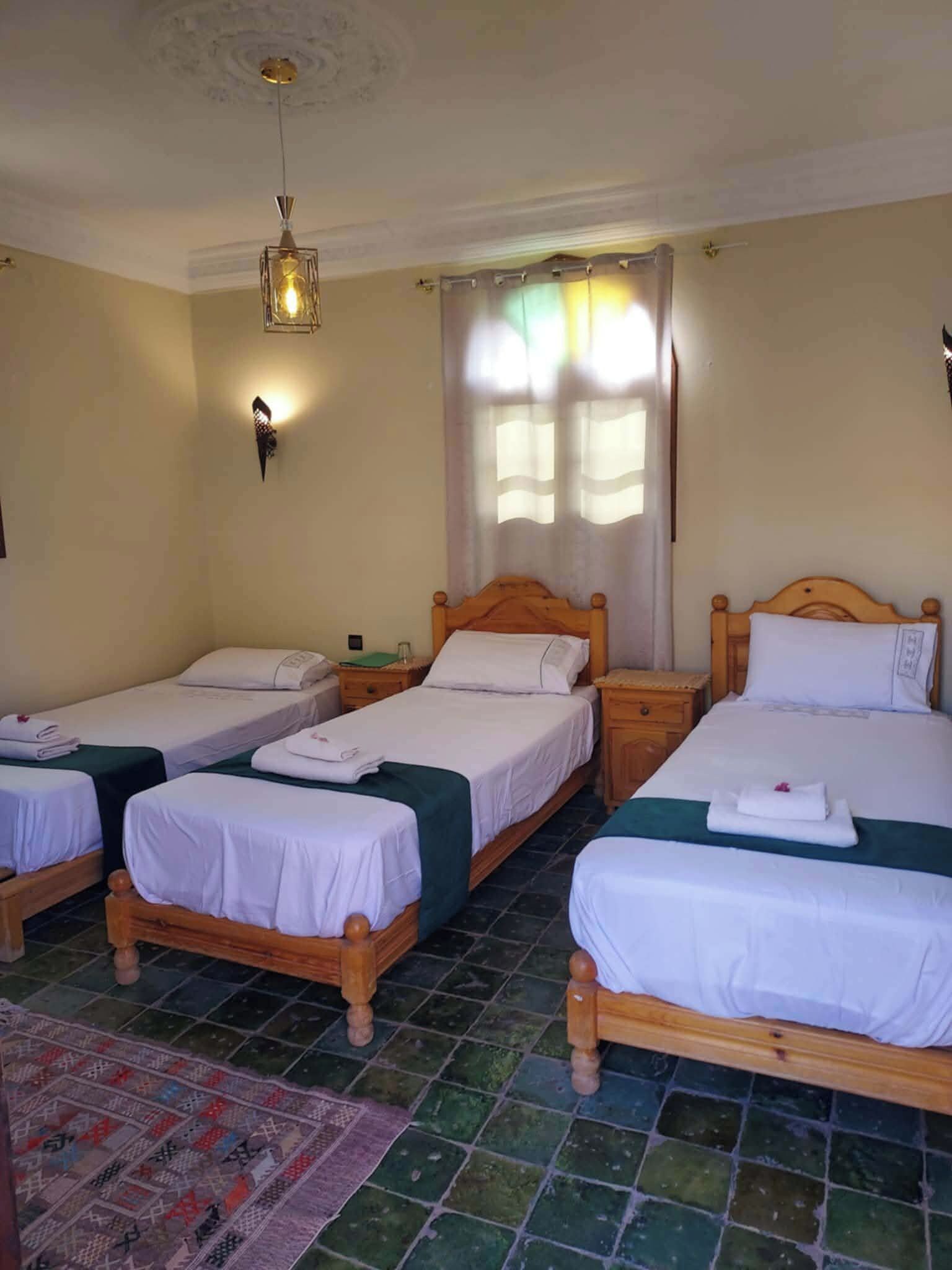 Une chambre d’hôtel avec trois lits en bois, un tapis à motifs, un luminaire fixé au mur et une fenêtre avec des rideaux.