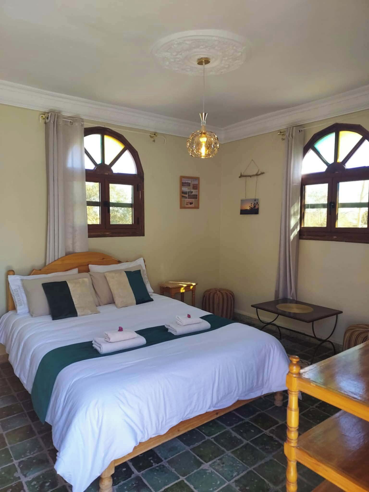 Una cama bien tendida con almohadas y toallas, una mesita pequeña y una silla en una habitación con dos ventanas y una lámpara en el techo.